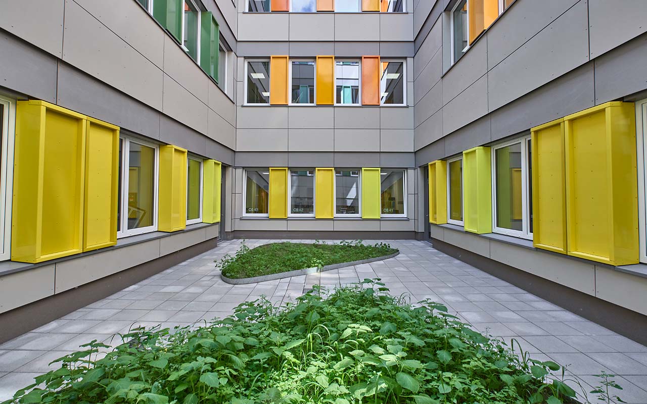Innenhof des Neubaus auf dem Schulcampus mit mehrgeschossigen Gebäudeflügeln, farbigen Fassadenelementen in Gelb und Grün sowie begrünten Pflanzflächen im zentralen Außenbereich.